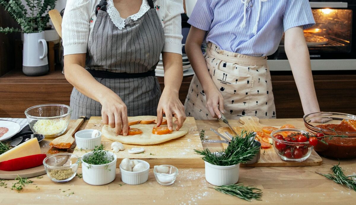 Crop unrecognizable females in aprons cooking pizza with tomatoes and cheese in kitchen together