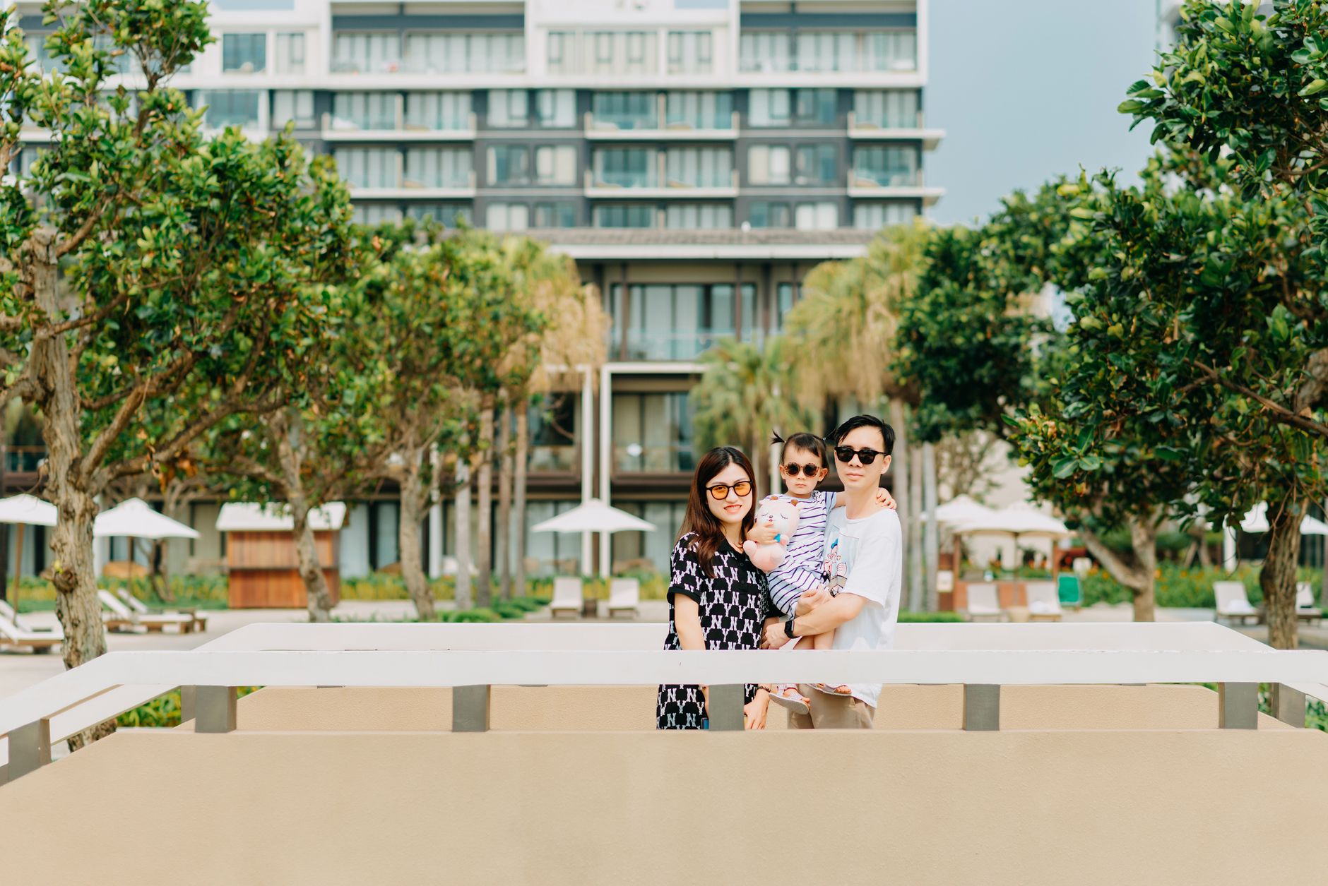 Positive young Asian couple with child wearing casual clothing and stylish sunglasses while standing among green tropical trees and looking at camera