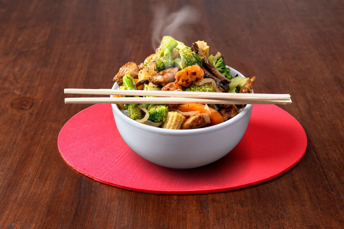 A steaming bowl of Asian stir fry with broccoli, carrots, mushrooms, and chopsticks on a red mat.