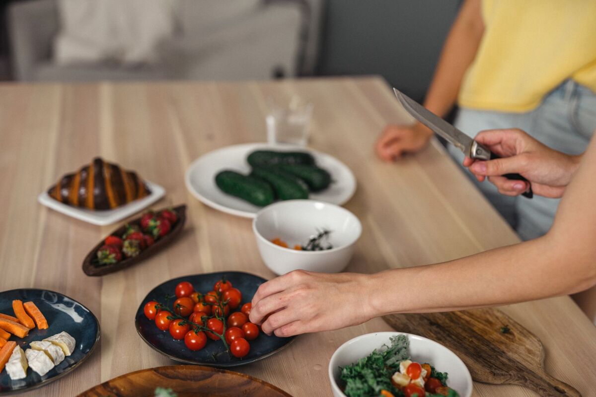 Hands slicing and arranging fresh vegetables and fruits on wooden table indoors.