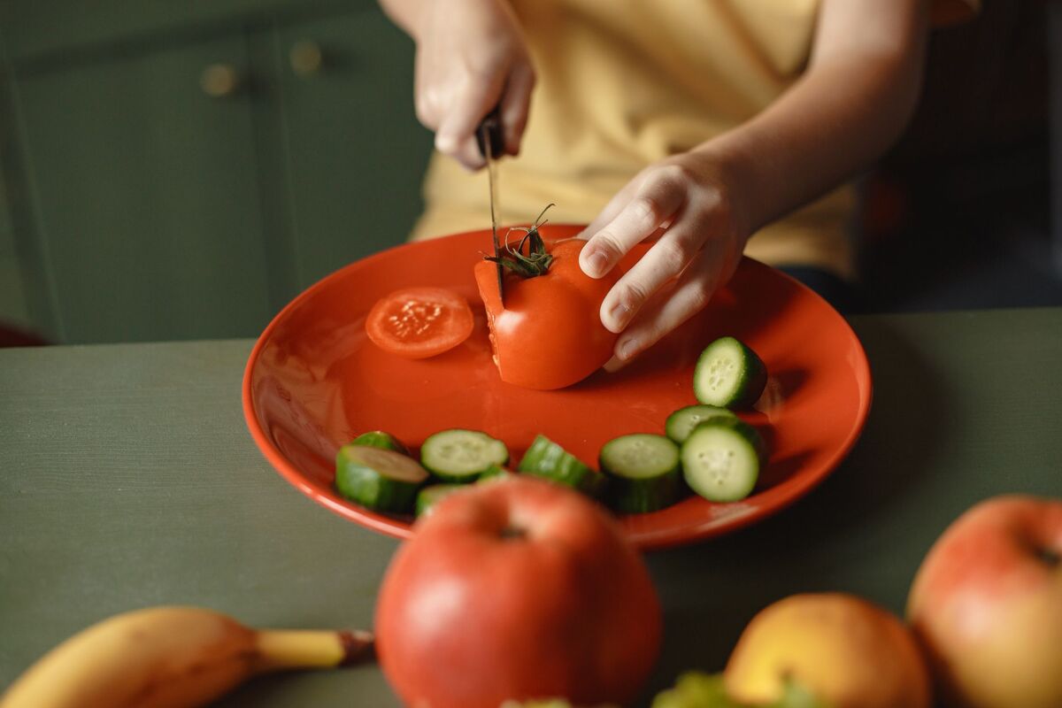 Unrecognizable kid cutting fresh tomato with knife on red ceramic dish with cucumber slices placed on table with assorted fruits while preparing healthy salad