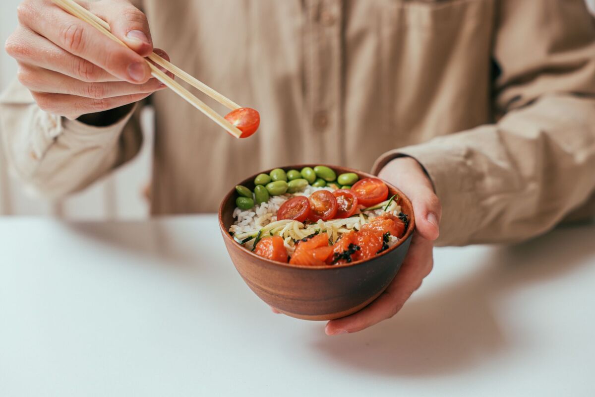A person holding a rice bowl with cherry tomatoes, edamame, and more, using chopsticks.