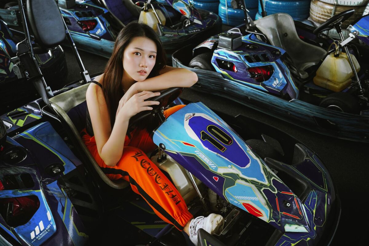 Portrait of a young woman sitting in a go-kart on an indoor track, with vibrant colors and a focused expression.