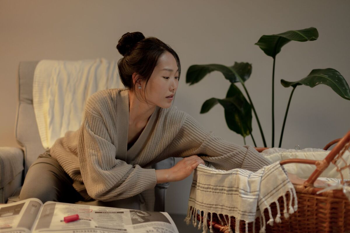 A thoughtful woman leans over a bassinet in a cozy home interior setting.