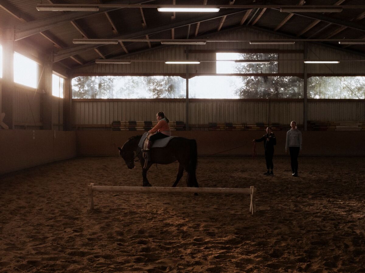 An indoor horse riding lesson with a child and two adults guiding the activity.