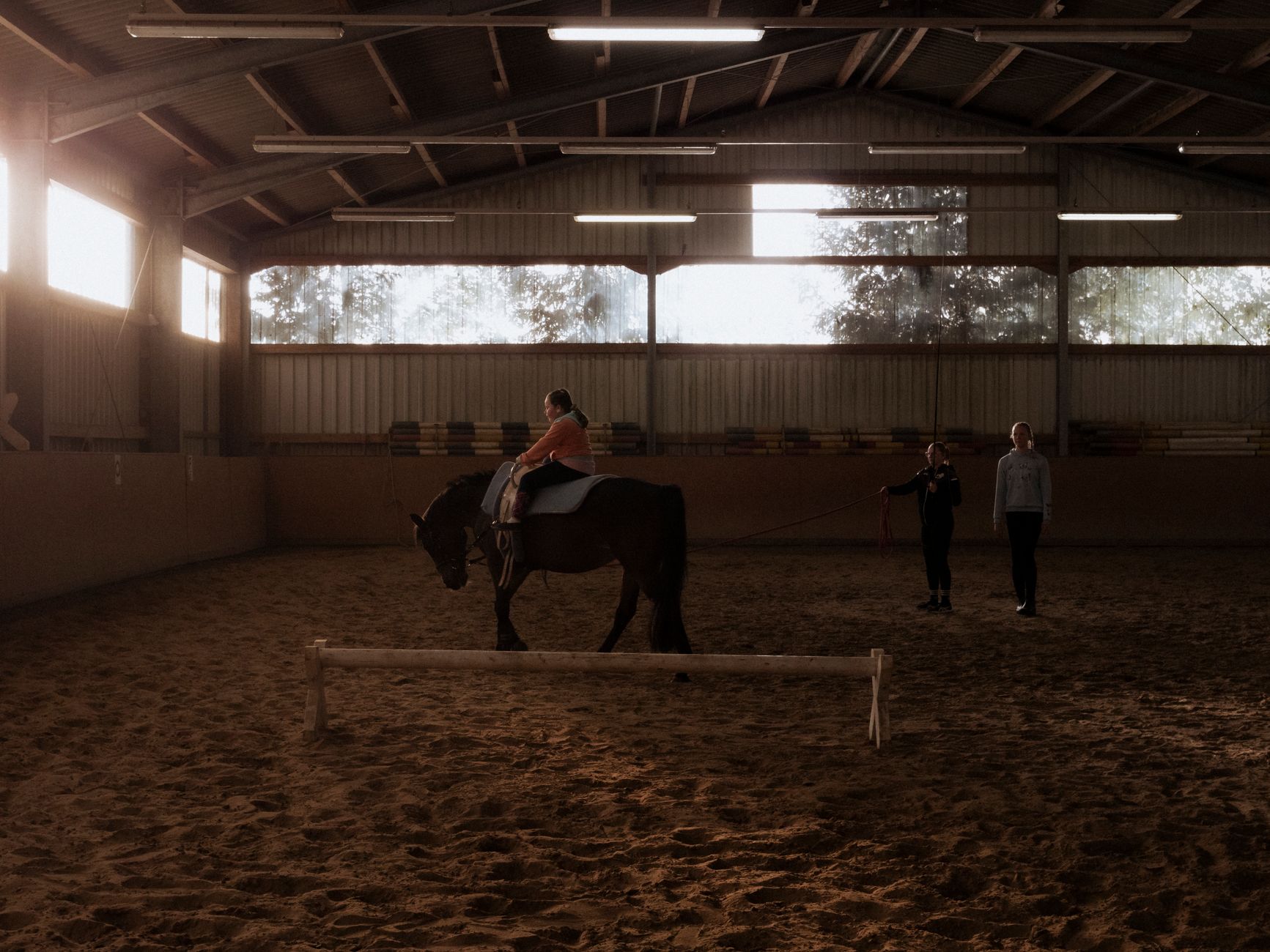 An indoor horse riding lesson with a child and two adults guiding the activity.