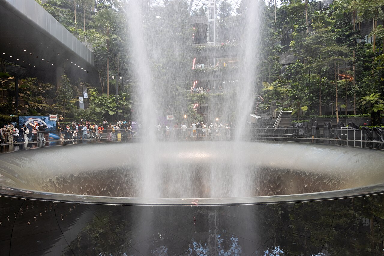 Ground-level view of the Rain Vortex waterfall splashing into its circular basin with visitors watching around the Forest Valley at Jewel Changi Airport