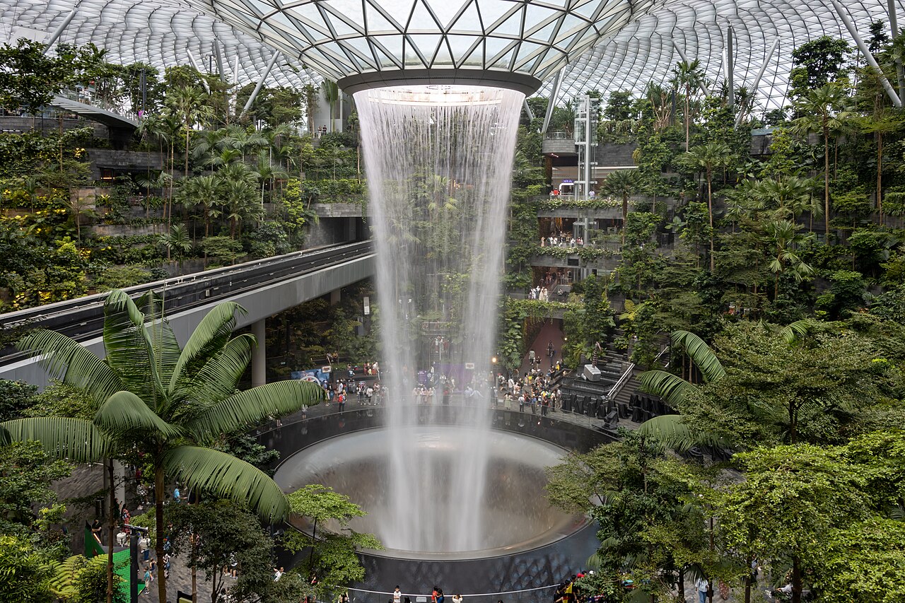 The column of the Rain Vortex waterfall framed by palms and lush greenery of the Shiseido Forest Valley at Jewel Changi Airport