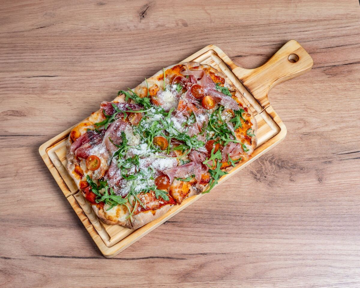 Delicious homemade Italian pizza topped with prosciutto, arugula, and cherry tomatoes on a wooden board.
