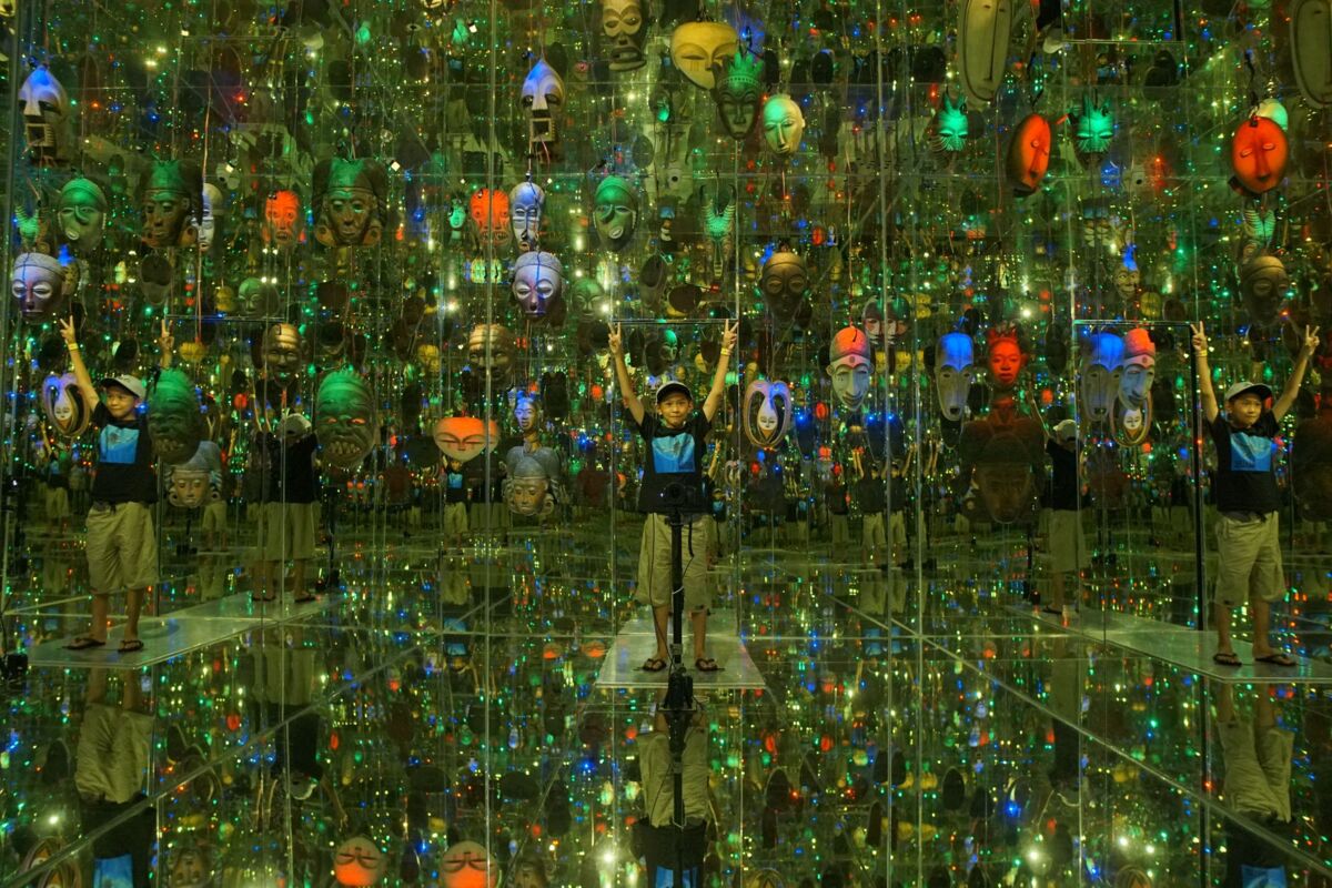 A child explores a vivid mirror room filled with colorful masks and lights.