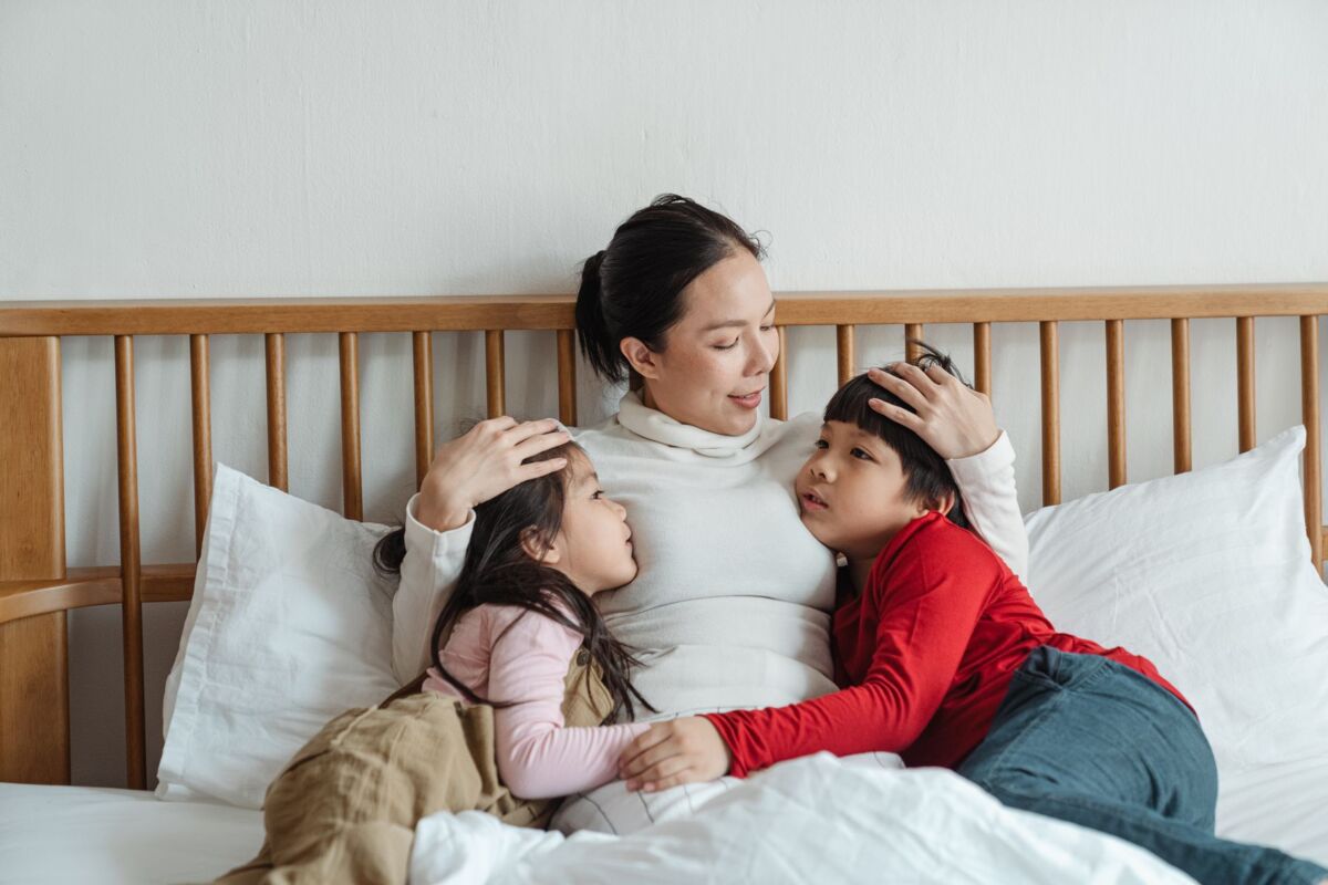 Positive ethnic mother patting little daughter and son on heads while children embracing woman lying on bed together