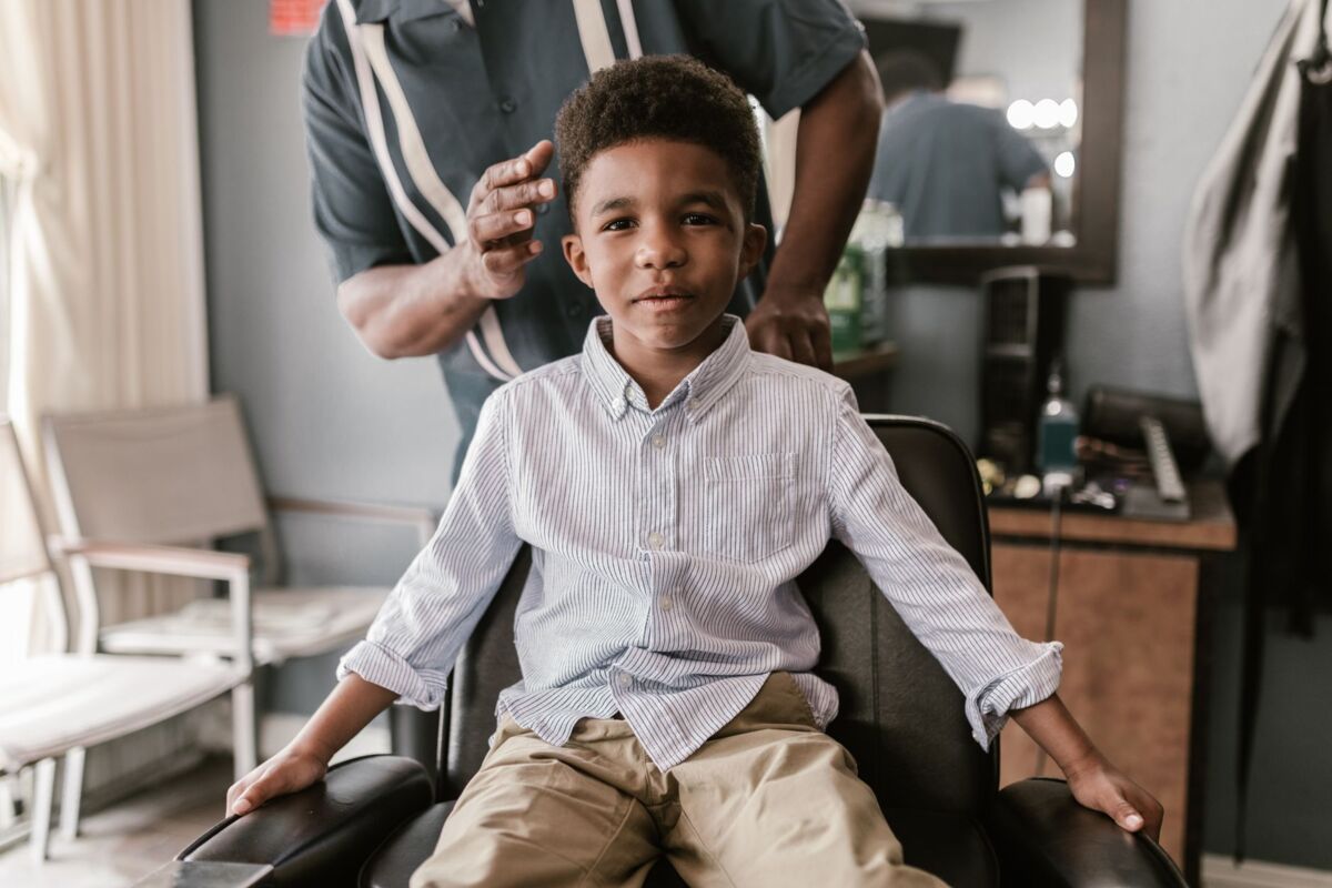A young boy in a striped shirt smiles while seated in a barbershop chair, anticipating a haircut.