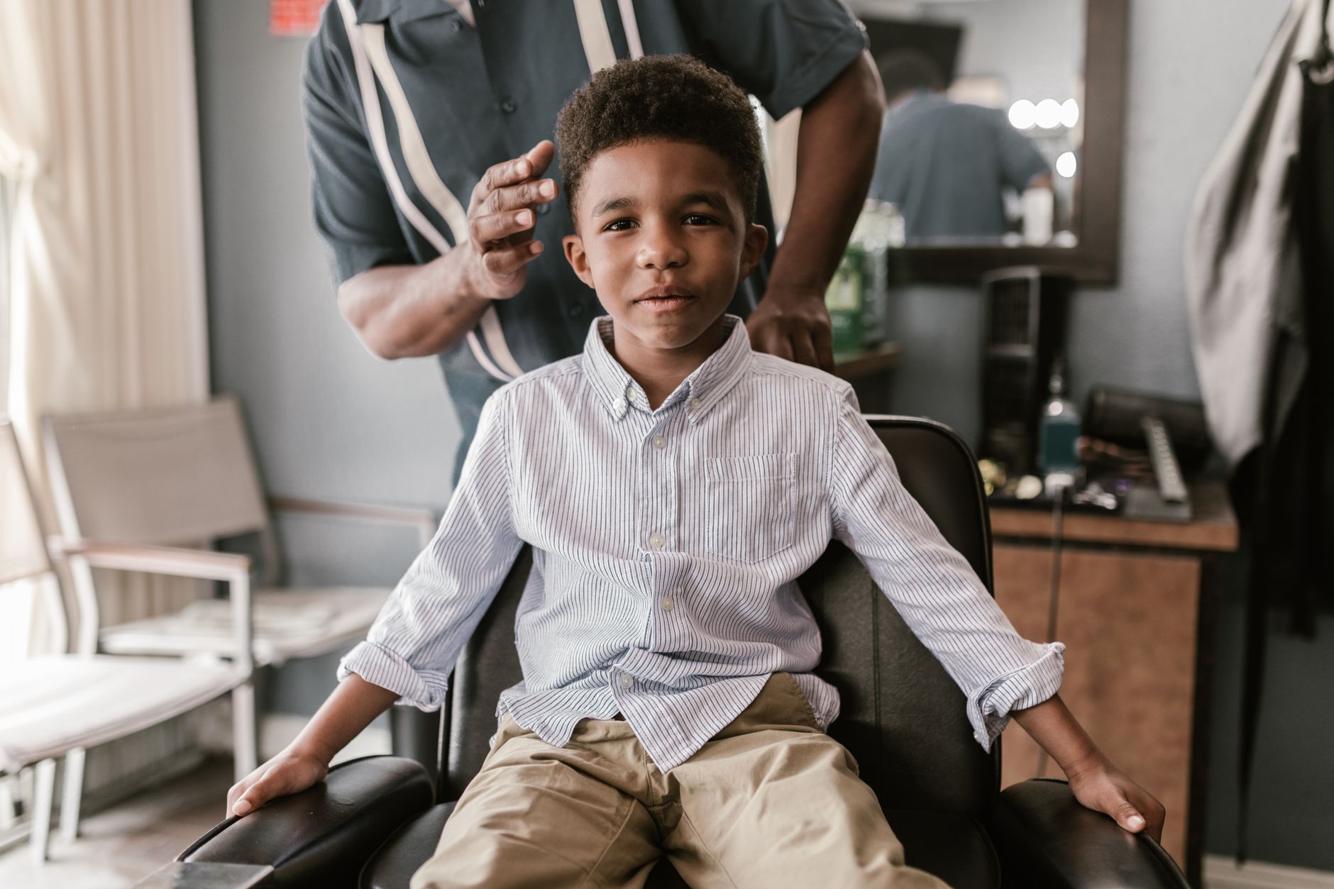 A young boy in a striped shirt smiles while seated in a barbershop chair, anticipating a haircut.