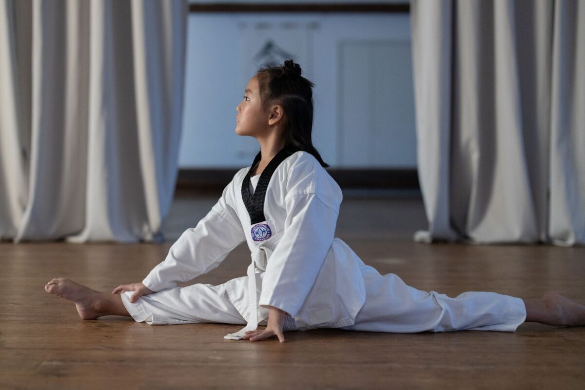 Asian girl in a dobok performing a taekwondo split indoors on a wooden surface.