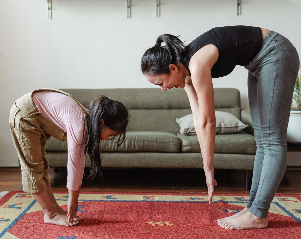 Side view of smiling young Asian woman stretching body with adorable daughter while standing against each other on floor carpet near cozy couch in modern living room