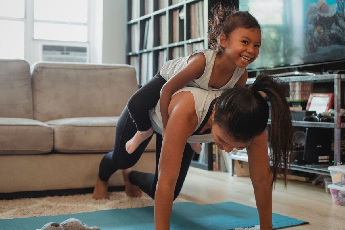 Fit Asian female in activewear piggybacking cute laughing daughter while performing exercise in light living room