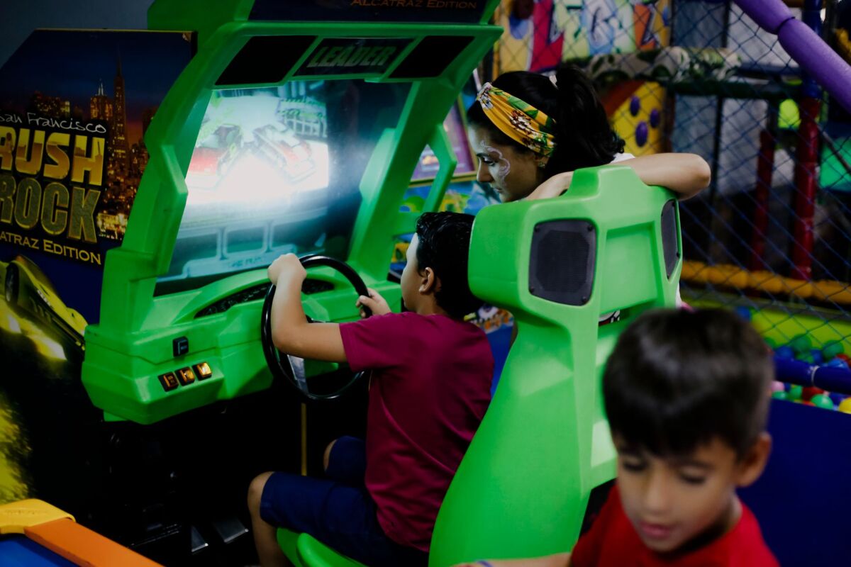 Unrecognizable child having fun with bright car racing simulator while turning steering wheel near serious ethnic woman and funny brother in shopping center