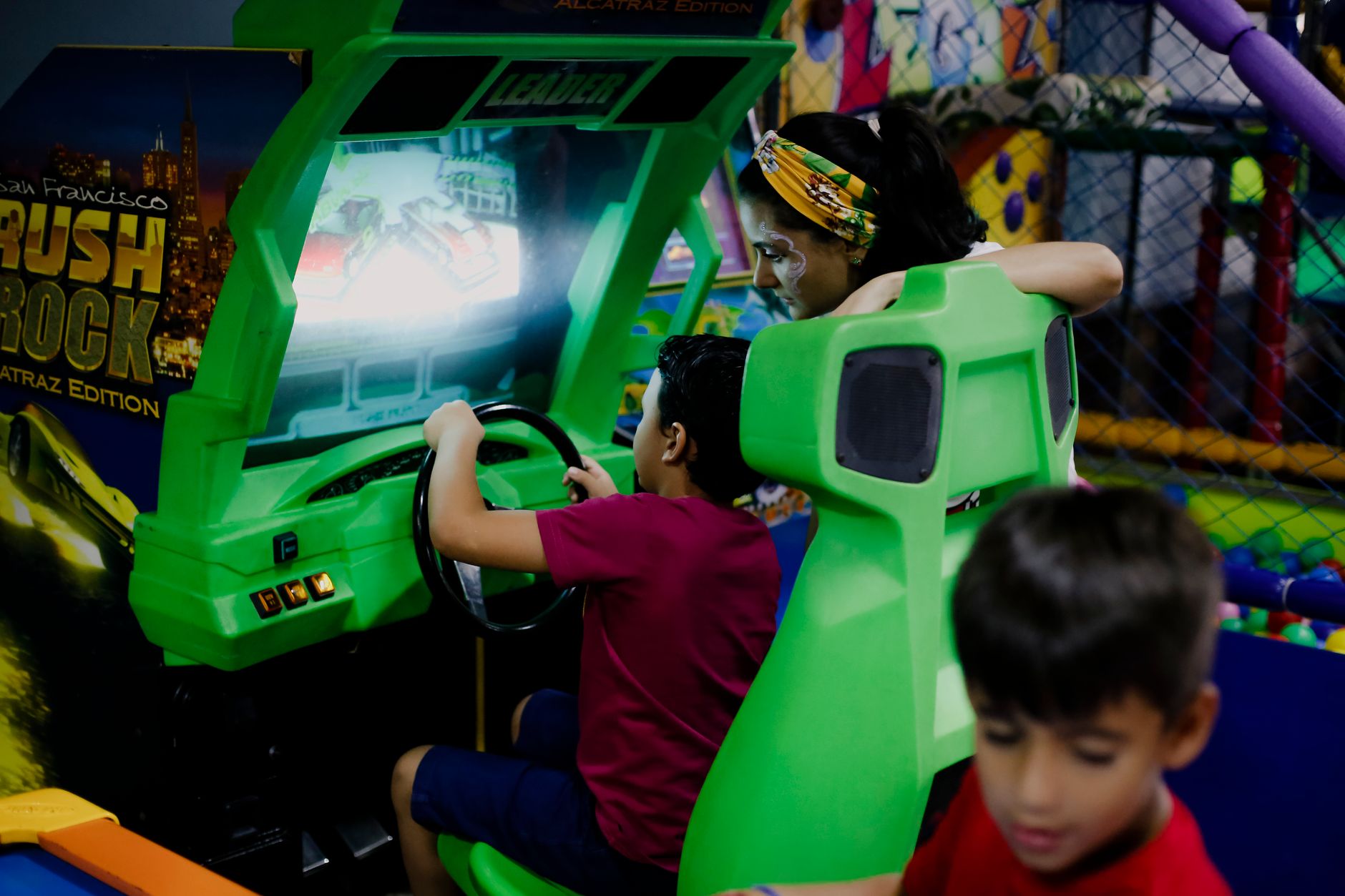 Unrecognizable child having fun with bright car racing simulator while turning steering wheel near serious ethnic woman and funny brother in shopping center