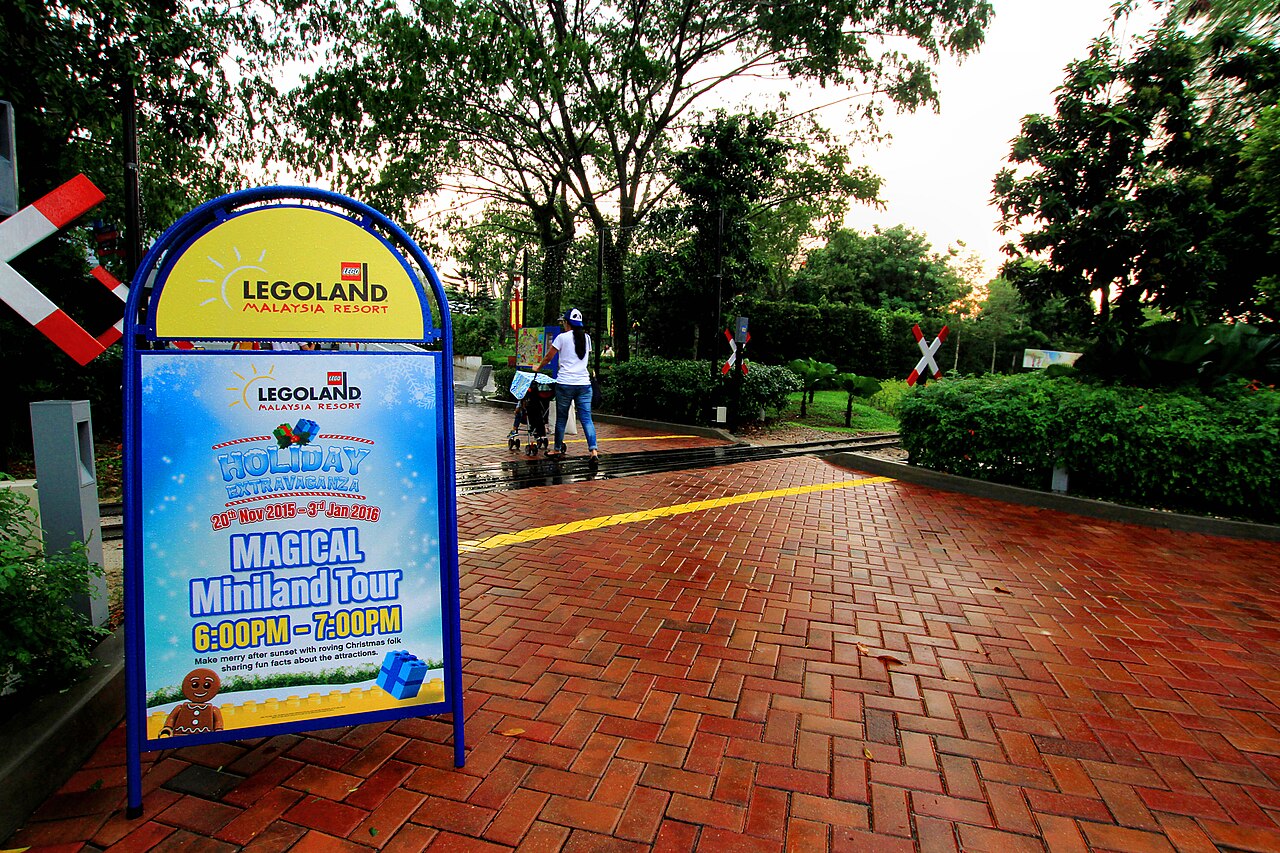 Legoland Malaysia Resort entrance signboard advertising a Miniland tour, with a parent pushing a stroller along a brick path