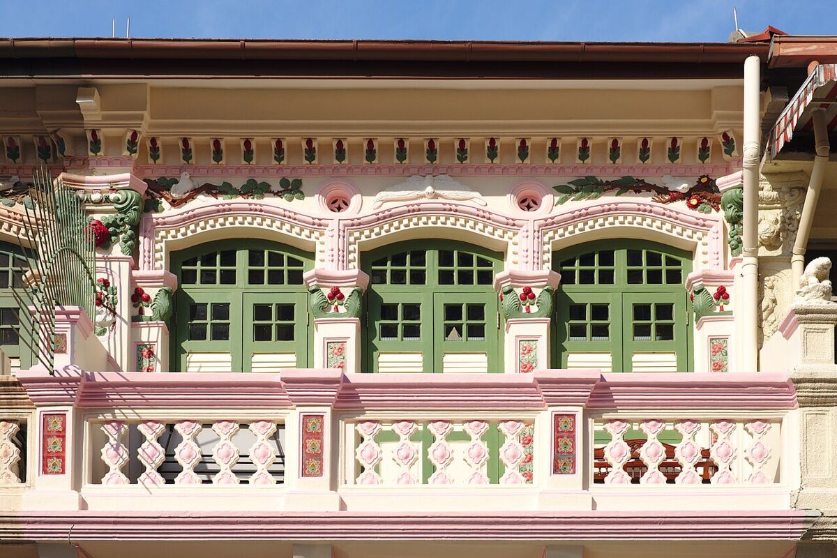 Row of colourful two-storey shophouses with shuttered windows and shop awnings facing a green park railing in Little India