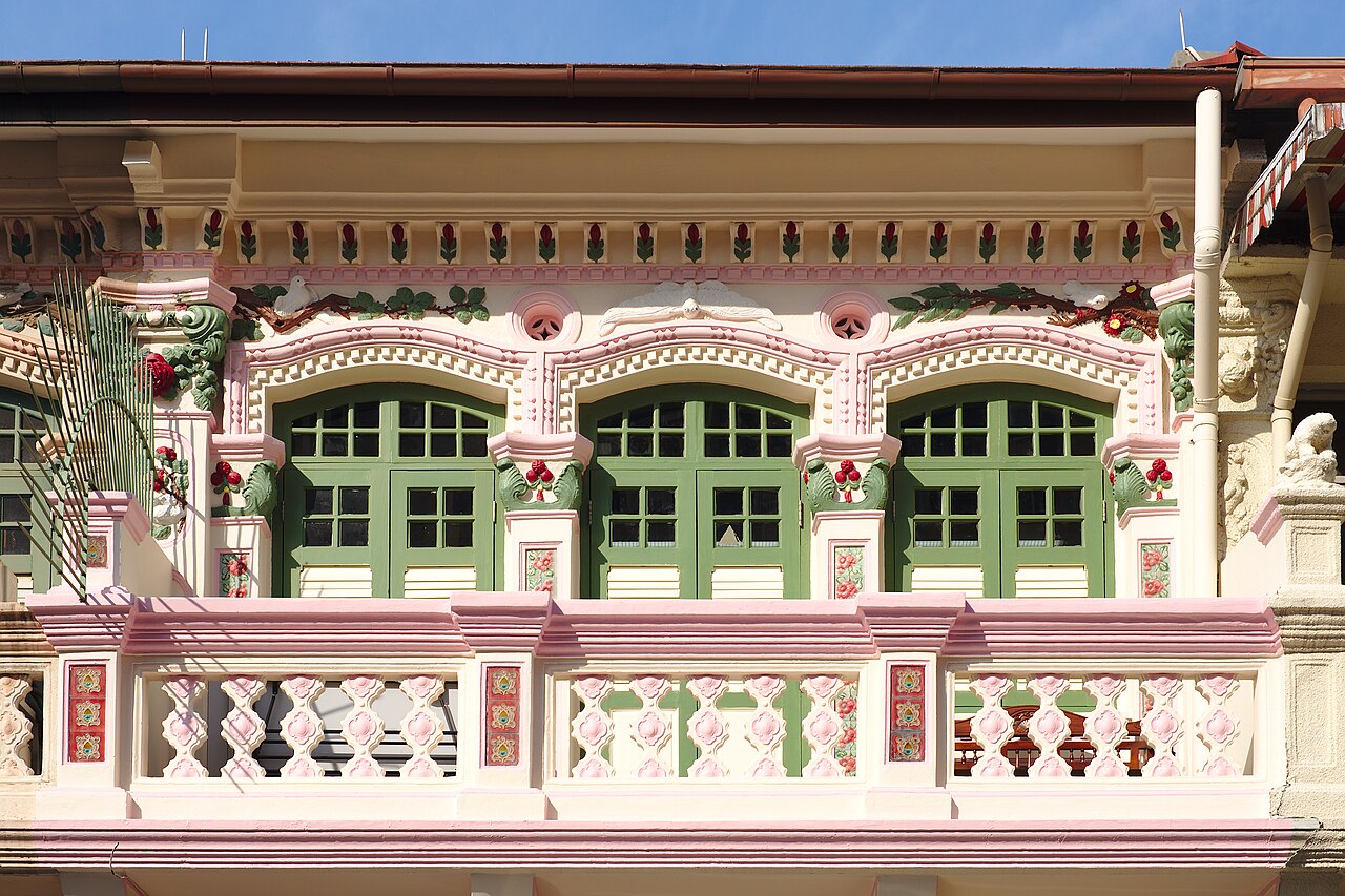 Row of colourful two-storey shophouses with shuttered windows and shop awnings facing a green park railing in Little India