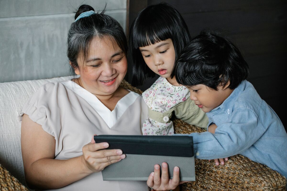 Positive Asian grandmother and kids looking at screen of tablet while resting at home