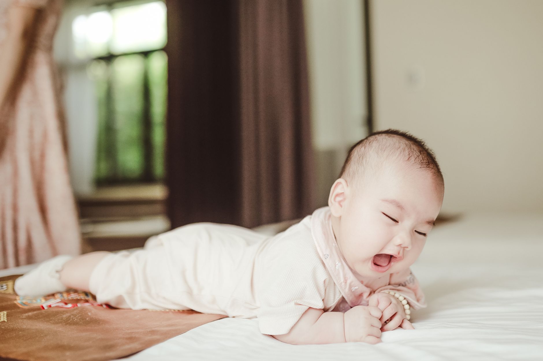 Adorable newborn baby in light pink bodysuit lying on belly on white soft coverlet on bed