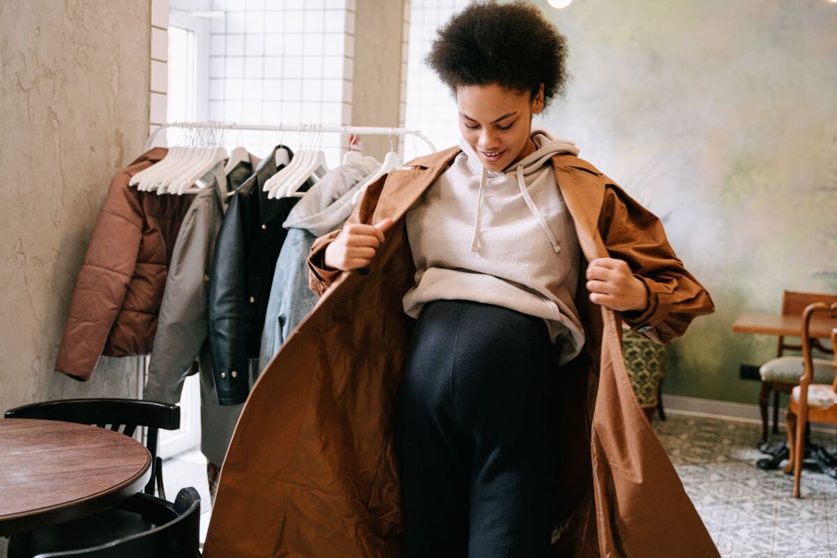 Pregnant woman in stylish coat adjusting outfit indoors near clothes rack.