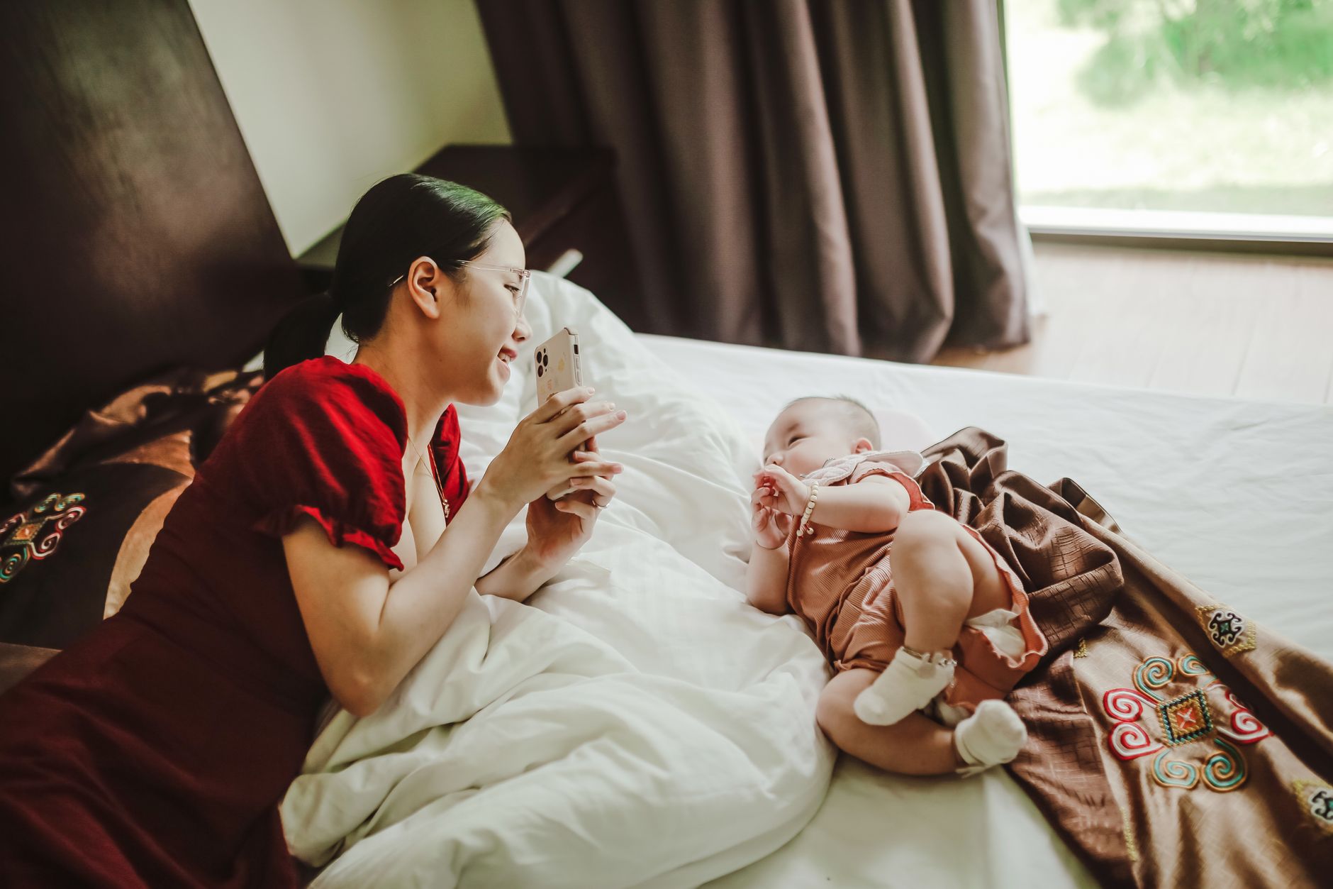Young Asian female mother showing smartphone to cute newborn baby lying on soft blanket on bed in light bedroom