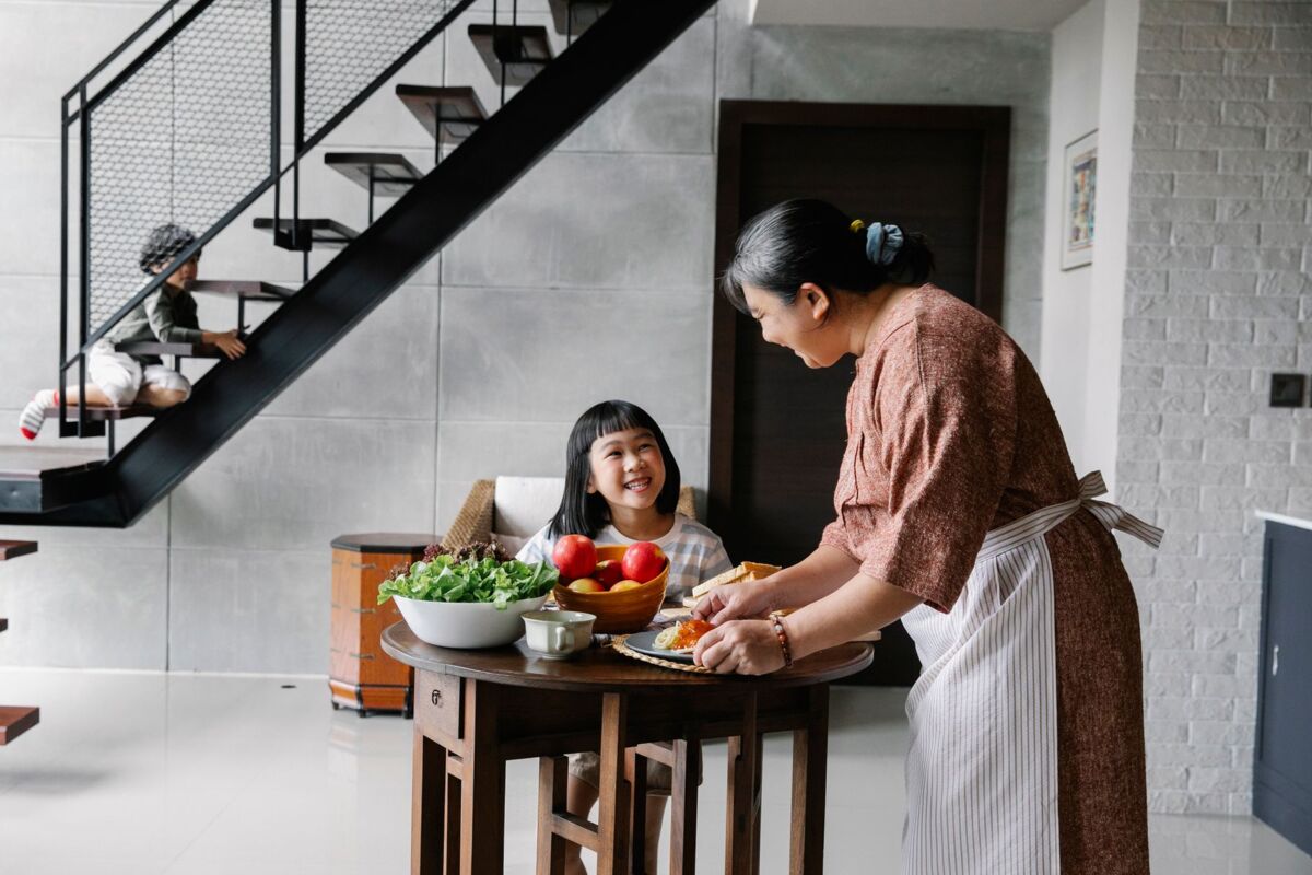 Side view of happy Asian grandmother serving delicious lunch with pasta and fresh vegetables for adorable little children while spending time together at home