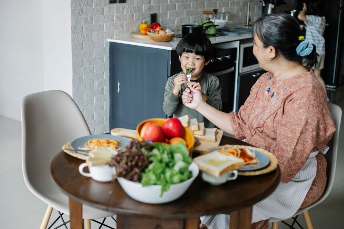Side view of positive senior Asian woman feeding cute little grandson during breakfast in modern kitchen in sunny morning.