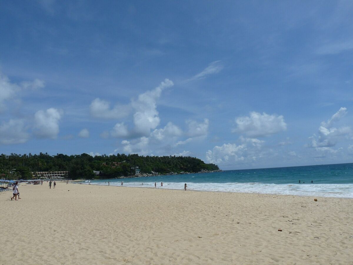 Wide sandy expanse of Karon Beach in Phuket, Thailand, with turquoise sea, gentle surf and a few people walking under a bright blue sky