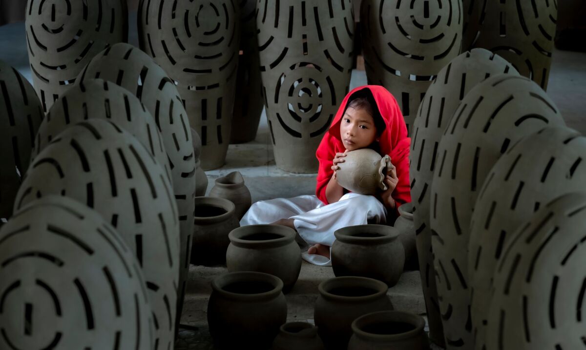 A young girl in a red hooded garment holds a clay pot in a pottery workshop, surrounded by ceramic vases.