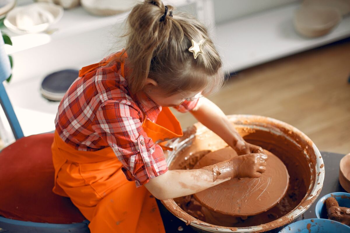 From above anonymous little girl in red check shirt and apron working with clay on pottery wheel while spending time in workshop