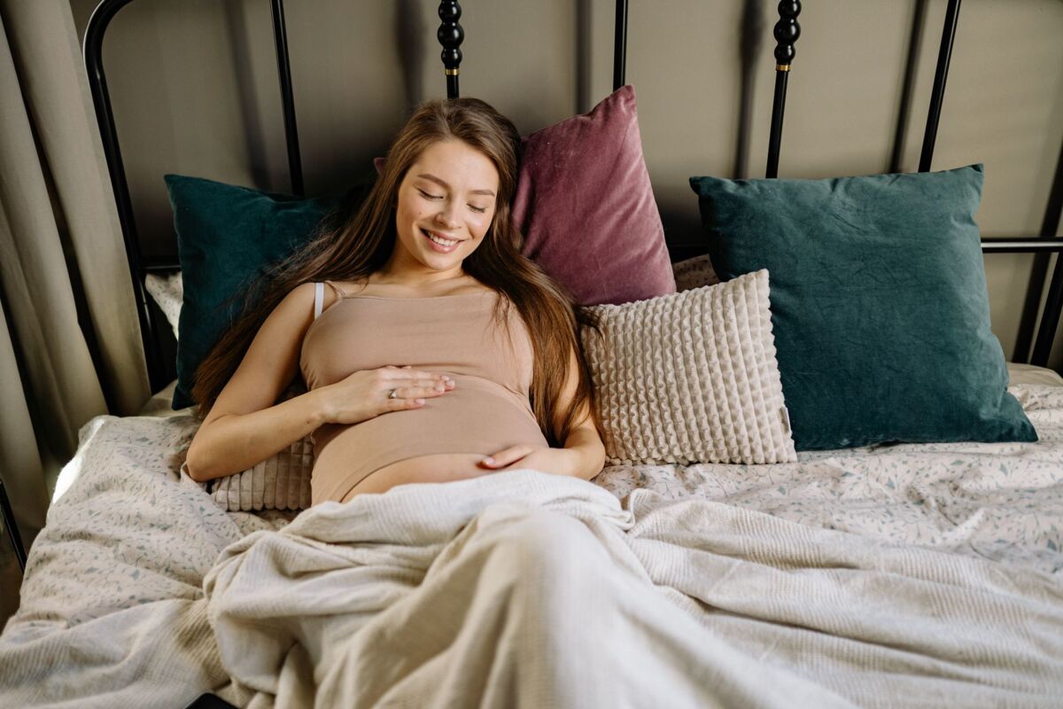 Smiling pregnant woman lying on a bed surrounded by colorful pillows, embracing motherhood.