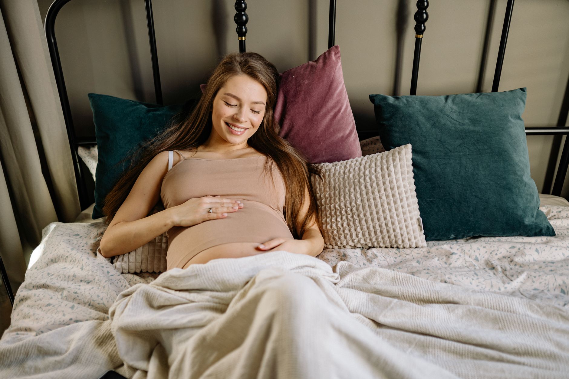 Smiling pregnant woman lying on a bed surrounded by colorful pillows, embracing motherhood.