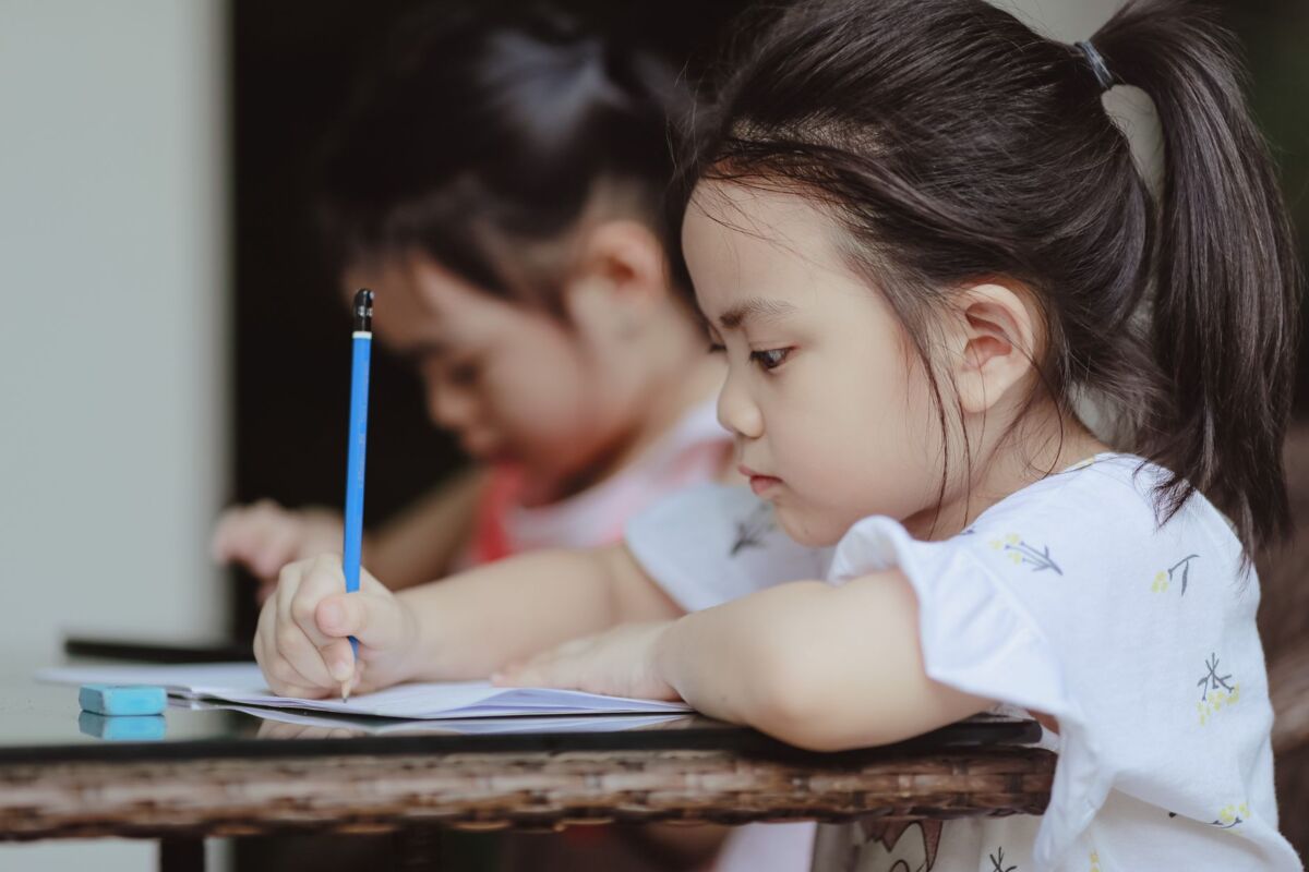 Adorable focused little Asian preschool girls sitting at table and doing tasks in copybooks in classroom