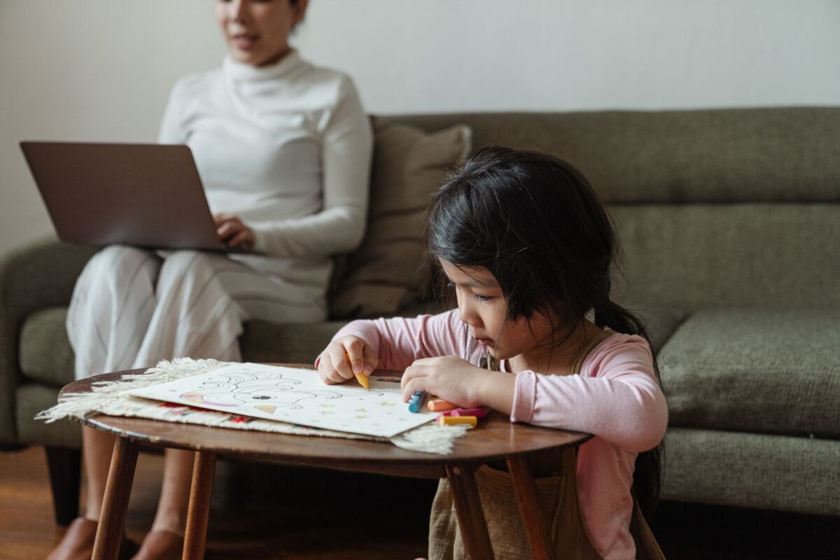 Focused ethnic little child in casual clothes sitting at coffee table on floor and coloring picture with colorful pencils against busy mother using laptop