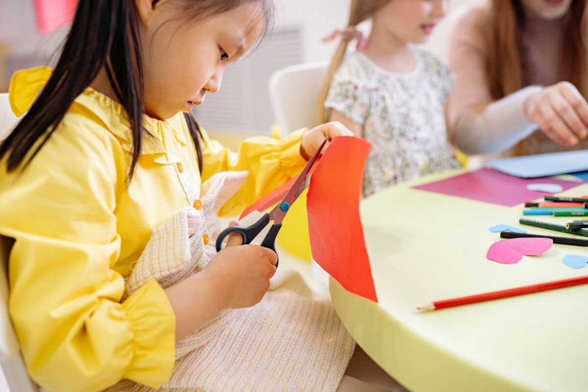 A young girl cuts colorful paper during an arts and crafts activity in a preschool classroom.