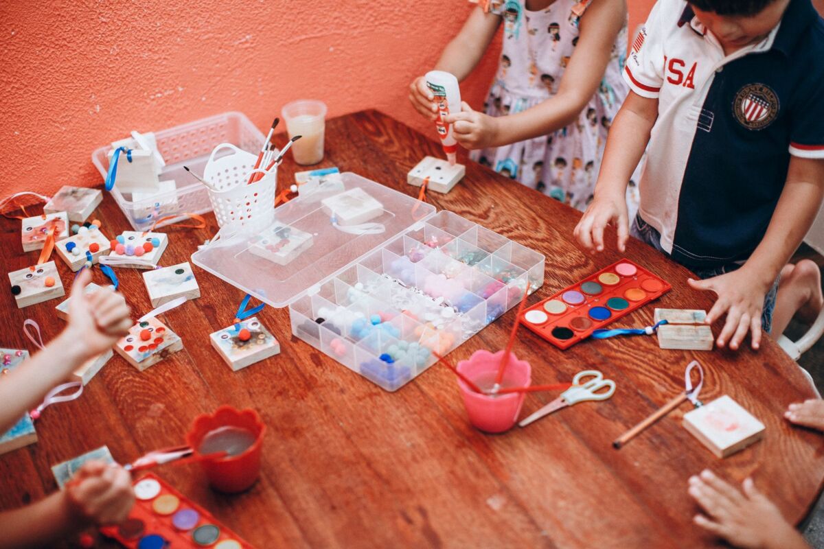 From above of crop anonymous children gathering around wooden table and painting with colorful watercolors