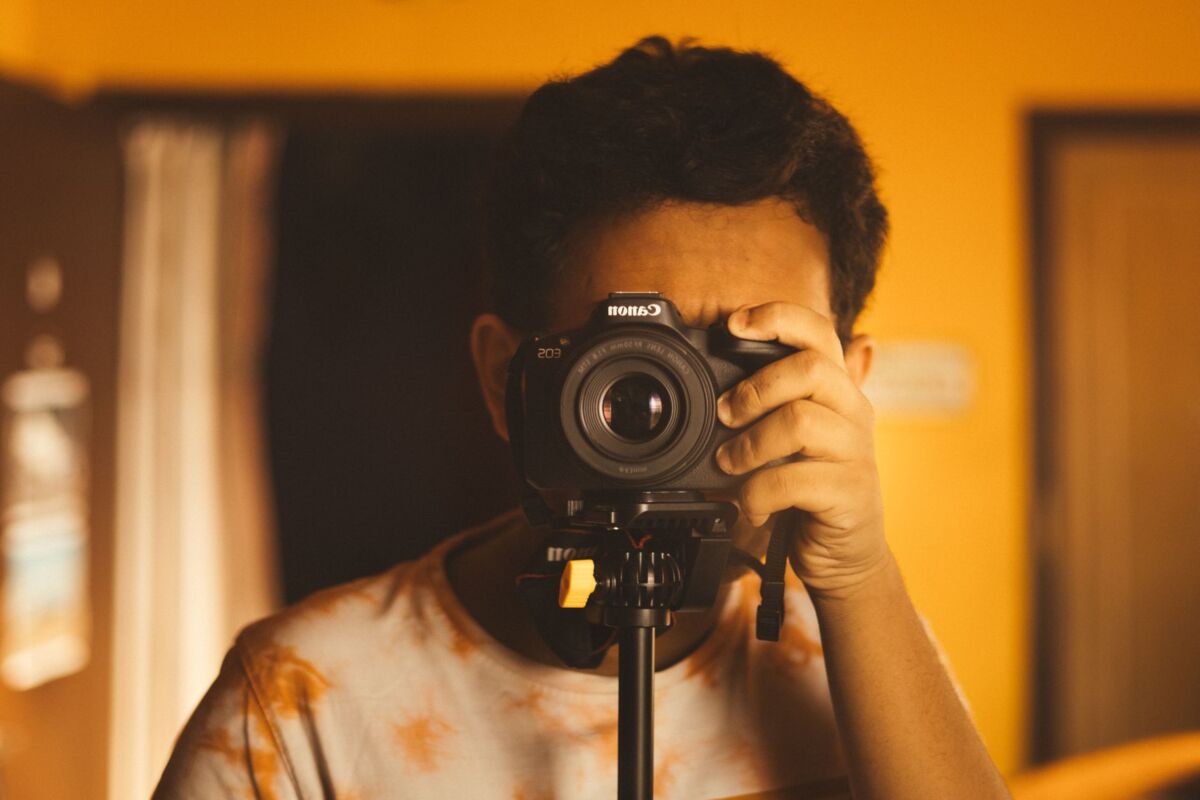 A young photographer captures a self-portrait indoors with a camera on a tripod, set against a warm, yellow background.