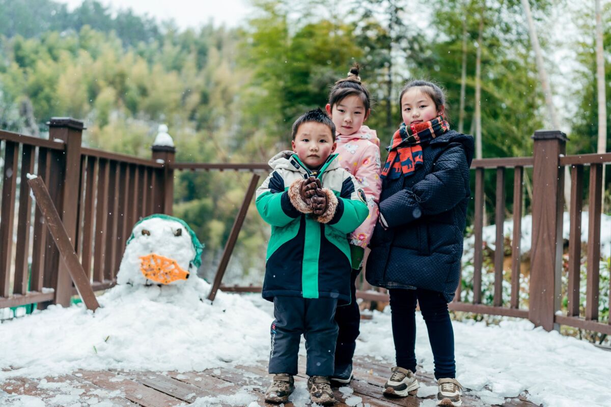 Three children enjoying winter outdoor play with a snowman in a scenic snowy setting.