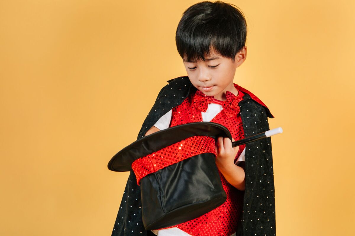 Cute small ethnic boy in costume of magician staring at hat on yellow background