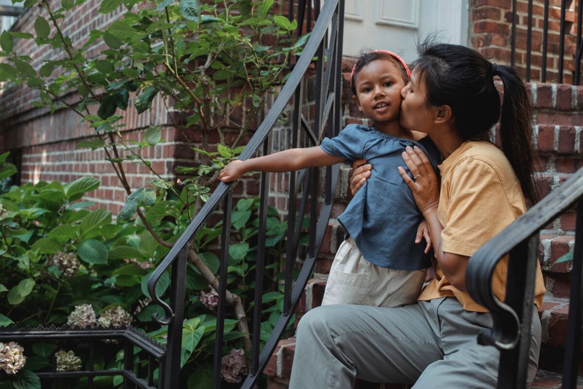 Tender ethnic mother hugging and kissing little girl while sitting on stairs of house with metal railing near green plants on terrace
