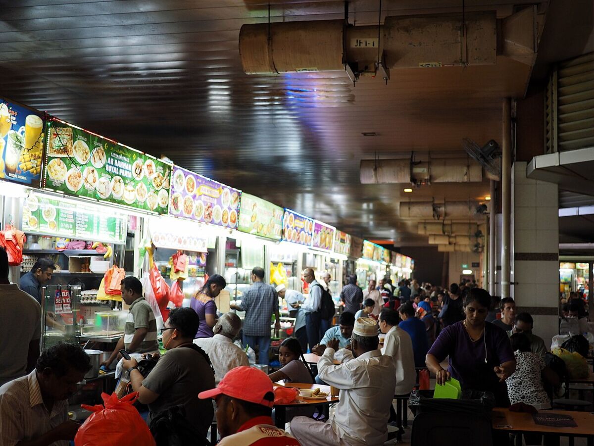 Busy Tekka Centre hawker food court with diners seated at tables beside food stalls