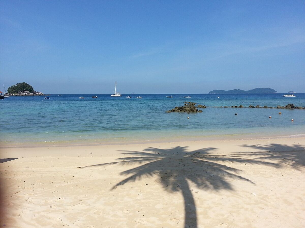 Palm tree shadow on white sand beach at Tioman Island with calm clear sea, boats and a sailing yacht offshore