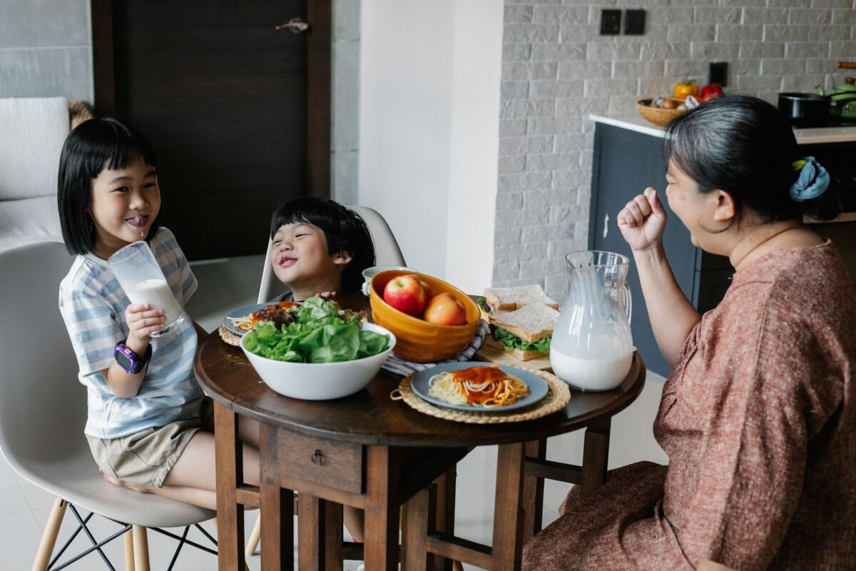 Cheerful Asian boy and girl sitting at served table with grandmother while having breakfast and drinking milk in morning on kitchen
