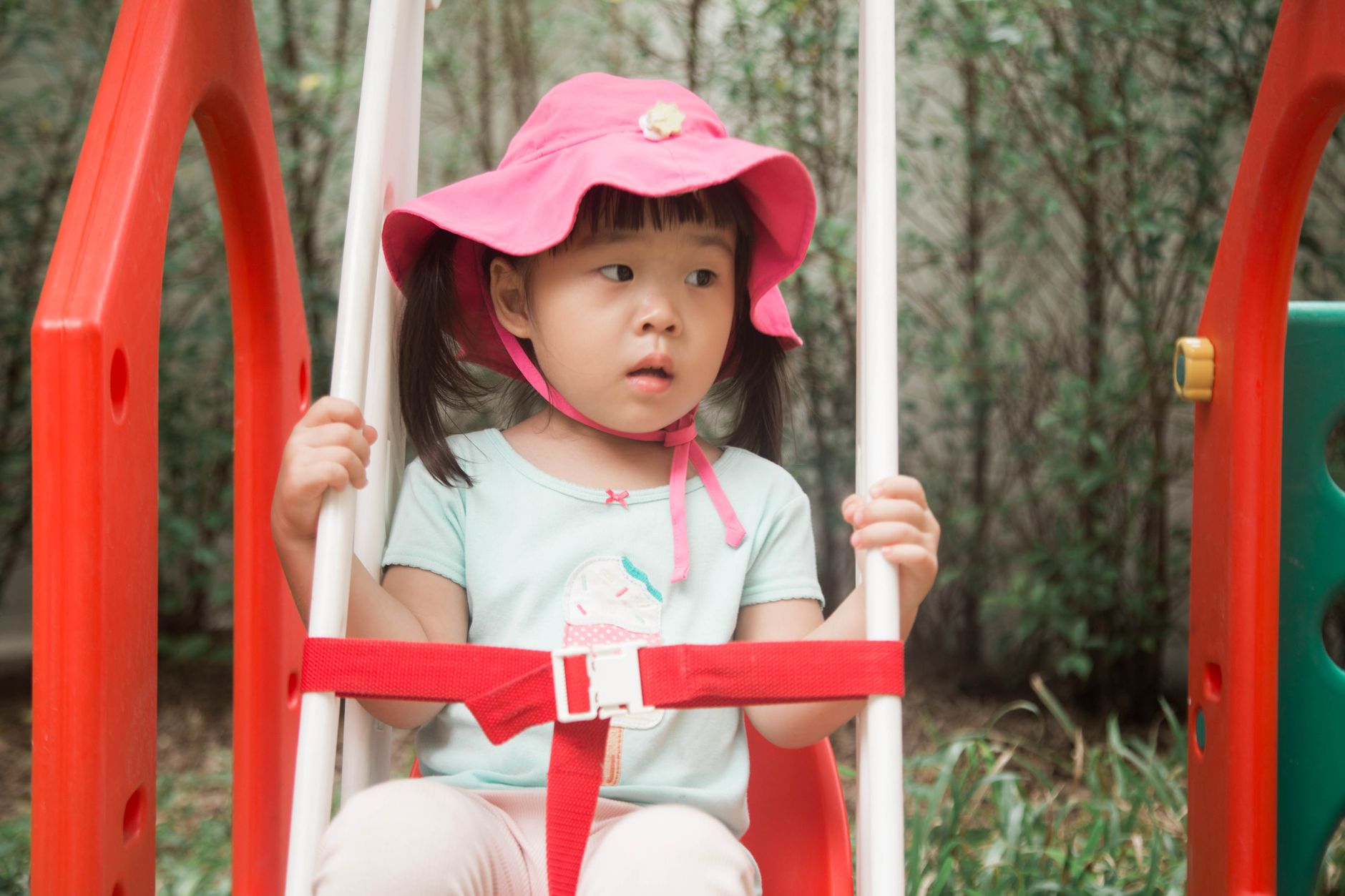 Cute Asian girl in a pink hat sitting on a swing at a playground.